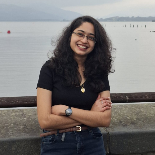 Irene is smiling and standing against a wall with her arms crossed. In the background there are some hills and a loch.